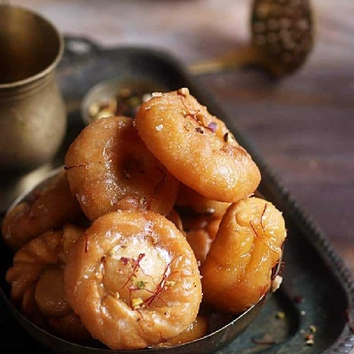 A close-up of Badusha, a popular Indian sweet, showing its round, flaky, and puffy shape with a golden-brown color. The sweet is made from maida flour, ghee, and yogurt, deep-fried to perfection, and then coated in a glossy, cardamom-flavored sugar syrup. Its crisp outer layer contrasts with the soft, melt-in-your-mouth interior, making it look irresistibly rich and delicious.