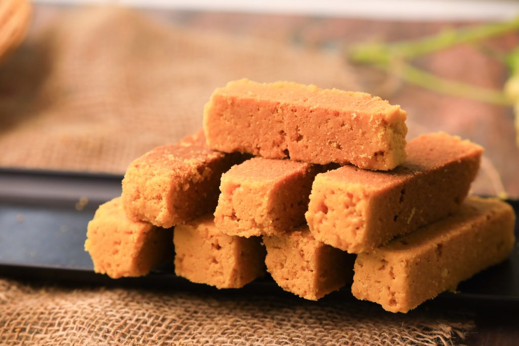 A close-up of Mysore Pak, a traditional South Indian sweet, cut into neat rectangular pieces. The sweet has a rich, golden-yellow color, with a smooth and slightly porous texture. Made from gram flour (besan), ghee, and sugar, the Mysore Pak looks soft and melts-in-the-mouth, with its glossy surface hinting at the richness of ghee. This classic dessert is a beloved treat, especially in South Indian households.