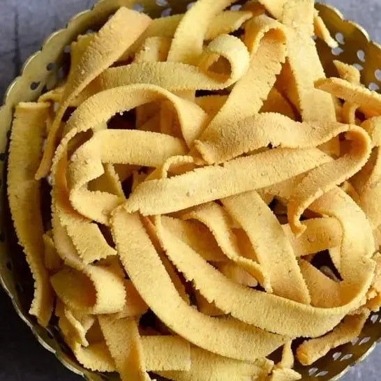 A pile of crispy, golden-brown ribbon pakkoda, a traditional South Indian snack, arranged on a plate. The snack is shaped into flat, wavy strips, showcasing its crunchy texture. Made from a spiced rice and gram flour dough, the pakkoda is deep-fried to perfection, giving it an appealing, crisp appearance. Its light and savory nature makes it an ideal snack for festive occasions or tea-time